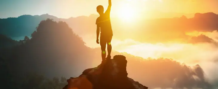 Silhouette of a person raising a fist in victory while standing on a mountain peak at sunrise.